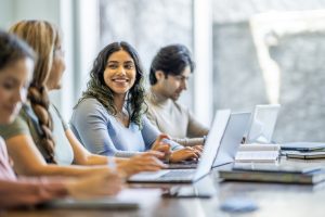 This image about A small group of four mature adult students sit together at a long desk as they study with one another. They have text books and laptops out in front of them as they take notes and talk amongst themselves.