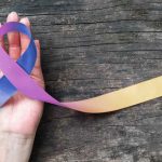 The image shows a hand holding a ribbon with three colours: blue, purple, and yellowish orange