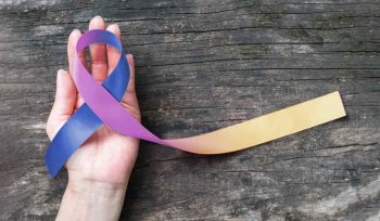 The image shows a hand holding a ribbon with three colours: blue, purple, and yellowish orange