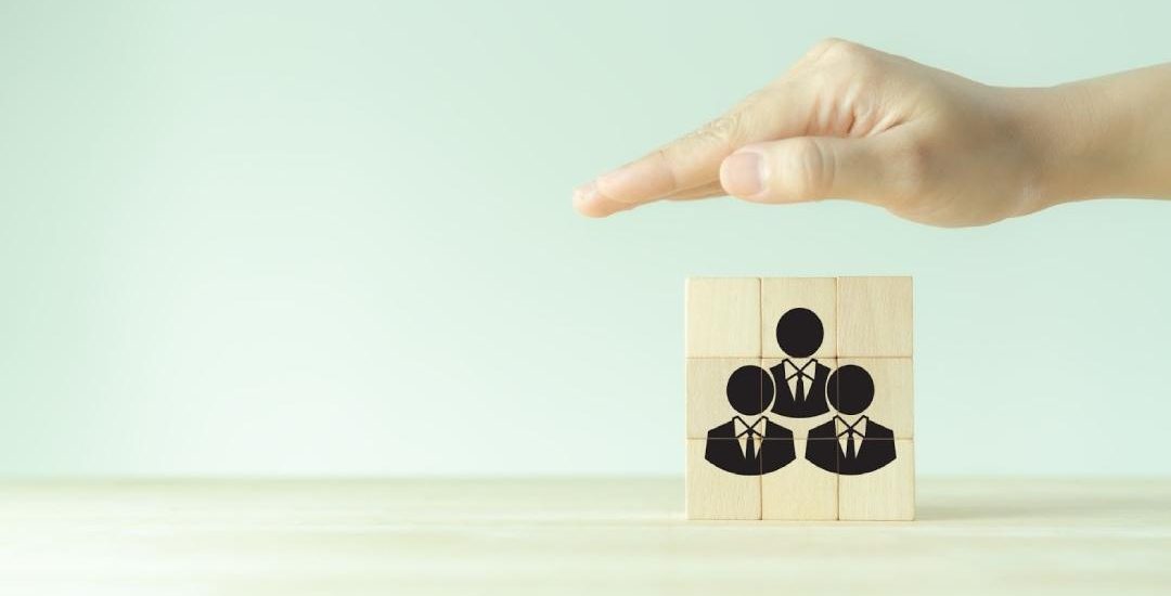 holi blog 1080x675 1 How to Structure an Employee Benefits Plan for Health Coverage A hand hovering over a stack of blocks, arranged to depict three people in suits