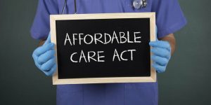 pros and cons of the affordable care act Can You Get Cancer Insurance for Employees in a Group Plan? The image shows a person wearing blue scrubs and gloves holding a small chalkboard with "AFFORDABLE CARE ACT" written on it